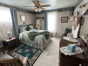 A bedroom with a bed, a dresser, a chair, a nightstand, a ceiling fan, and a window with curtains. - The Alma House Inn (Silverton)