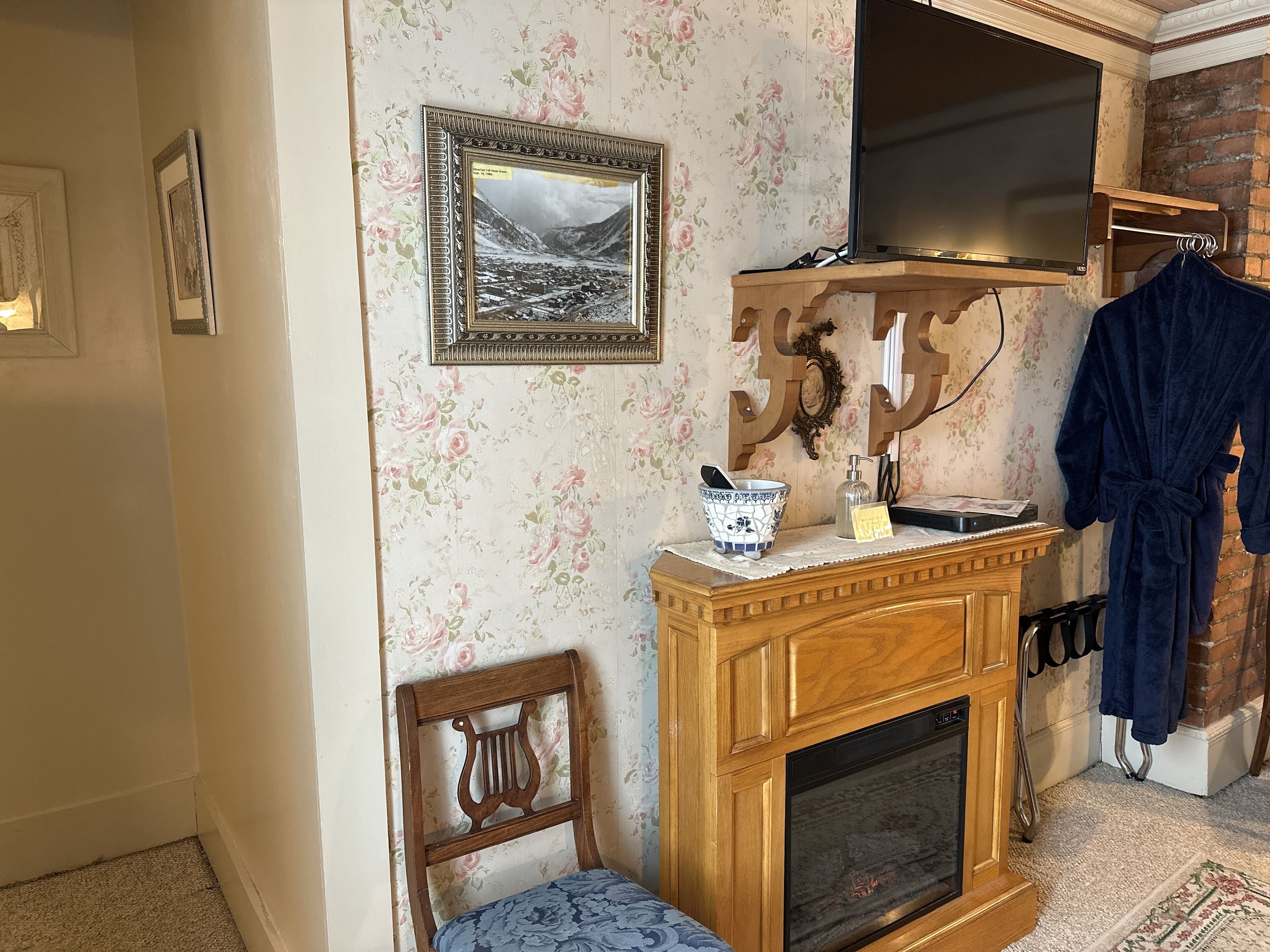 A room with a fireplace, a wooden chair, a blue patterned sofa, a wooden cabinet, and a blue robe hanging on a rack.
