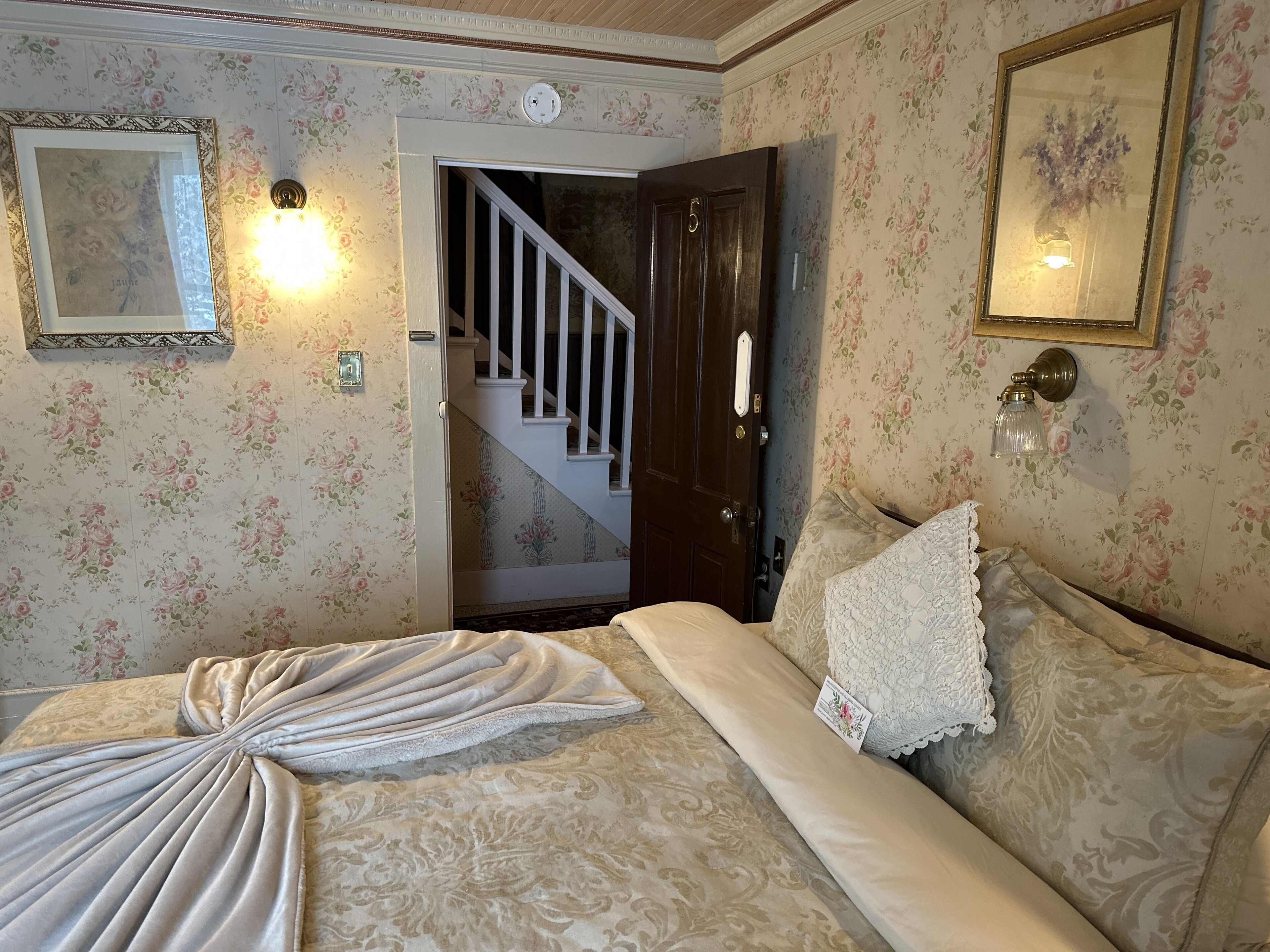 Un dormitorio con papel tapiz floral, una cama con manta decorativa, un cuadro enmarcado y una escalera.
