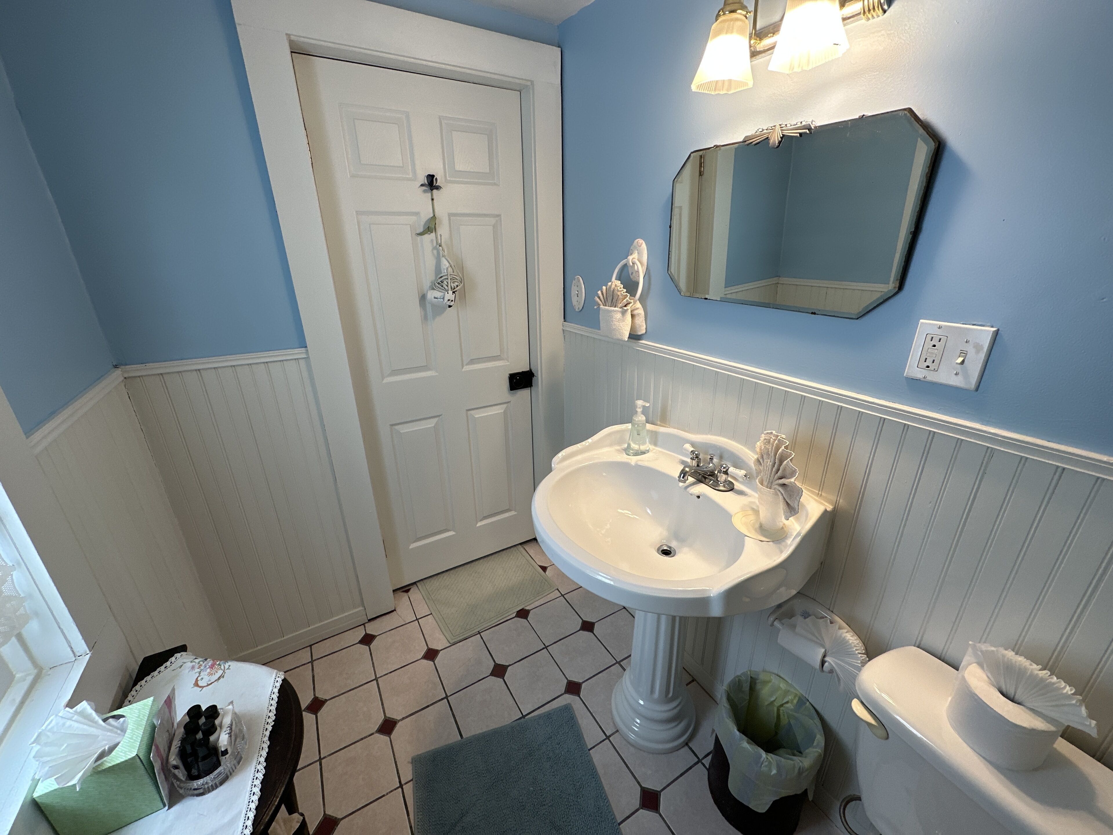 A bathroom with a white sink, a mirror, and a toilet. There is a door with a towel hanging on it, and a trash can is visible.