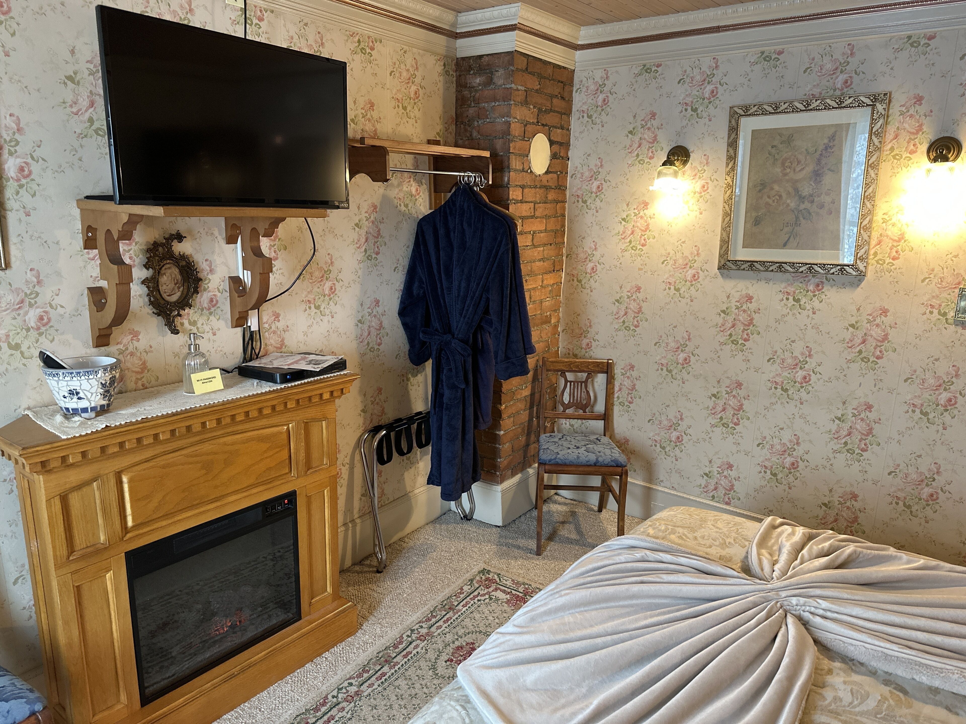 A bedroom with a fireplace, a television, a bathrobe, a chair, and a bed with a draped cover.
