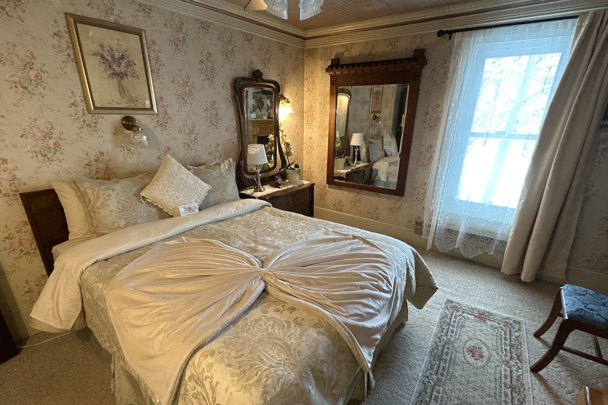 A bedroom with floral wallpaper, a large bed with a decorative headboard, a nightstand with a lamp, a mirror, and a window with curtains.