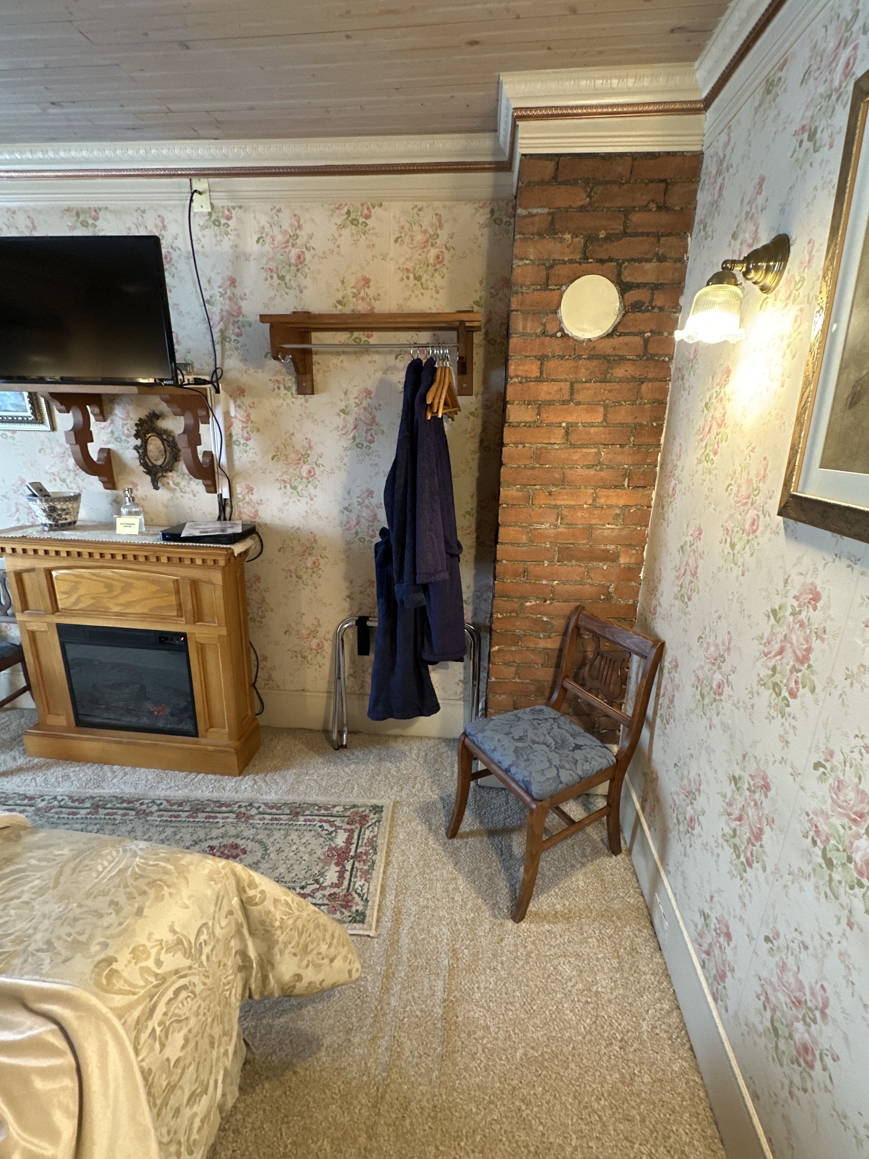 A room with a wooden cabinet, a television, a brick wall, a floral wallpaper, and a chair with a cushion.