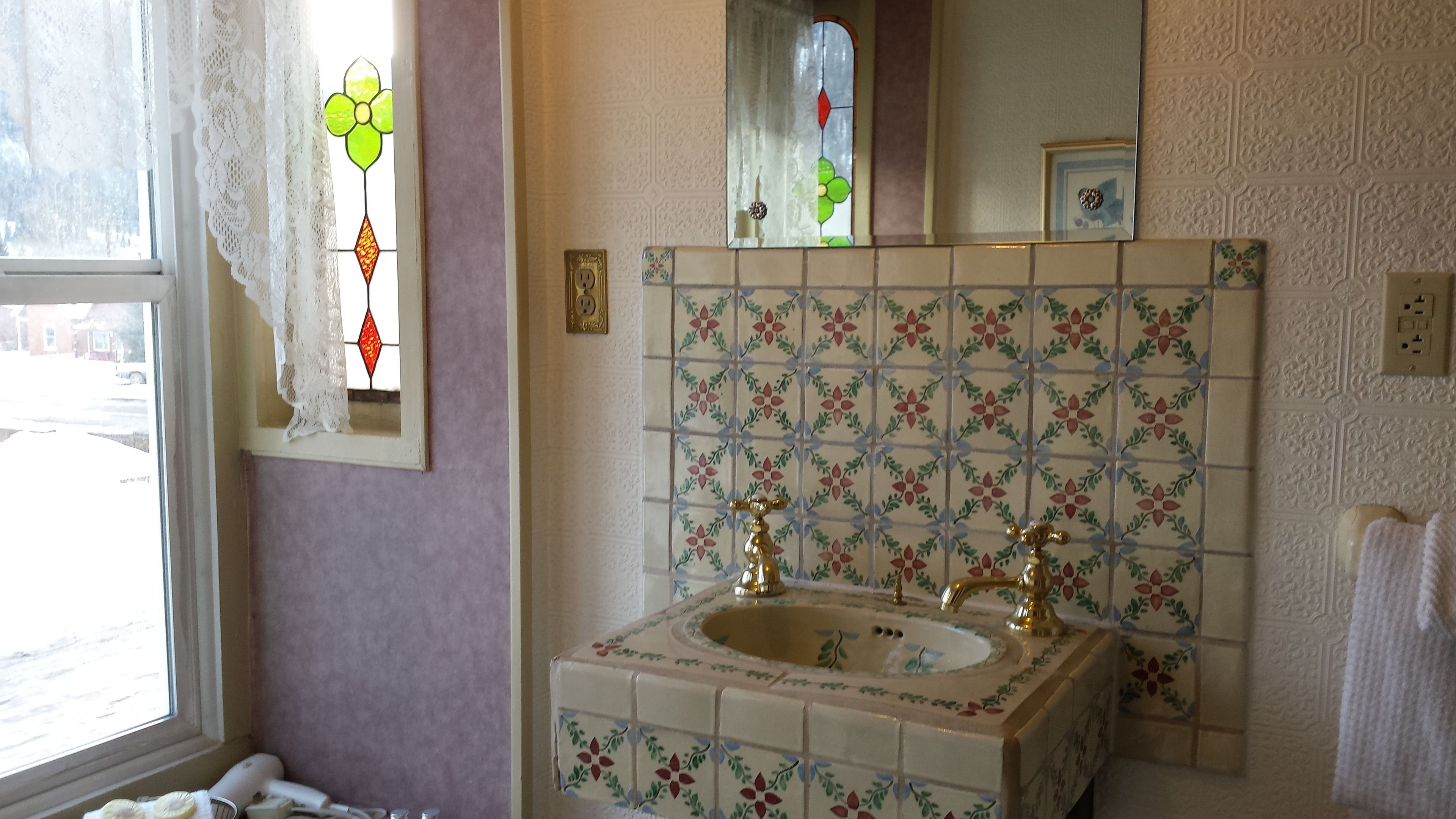 A bathroom with a floral tile backsplash, a sink with two gold faucets, and a window with lace curtains.