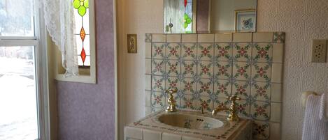 A bathroom with a floral tile backsplash, a sink with two gold faucets, and a window with lace curtains.