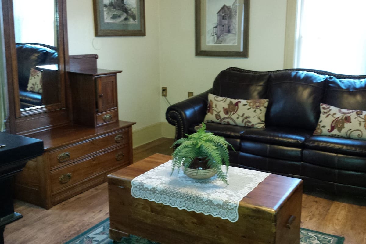 A living room with a dark brown leather sofa, a wooden coffee table with a lace tablecloth, a wooden cabinet, and a mirror.