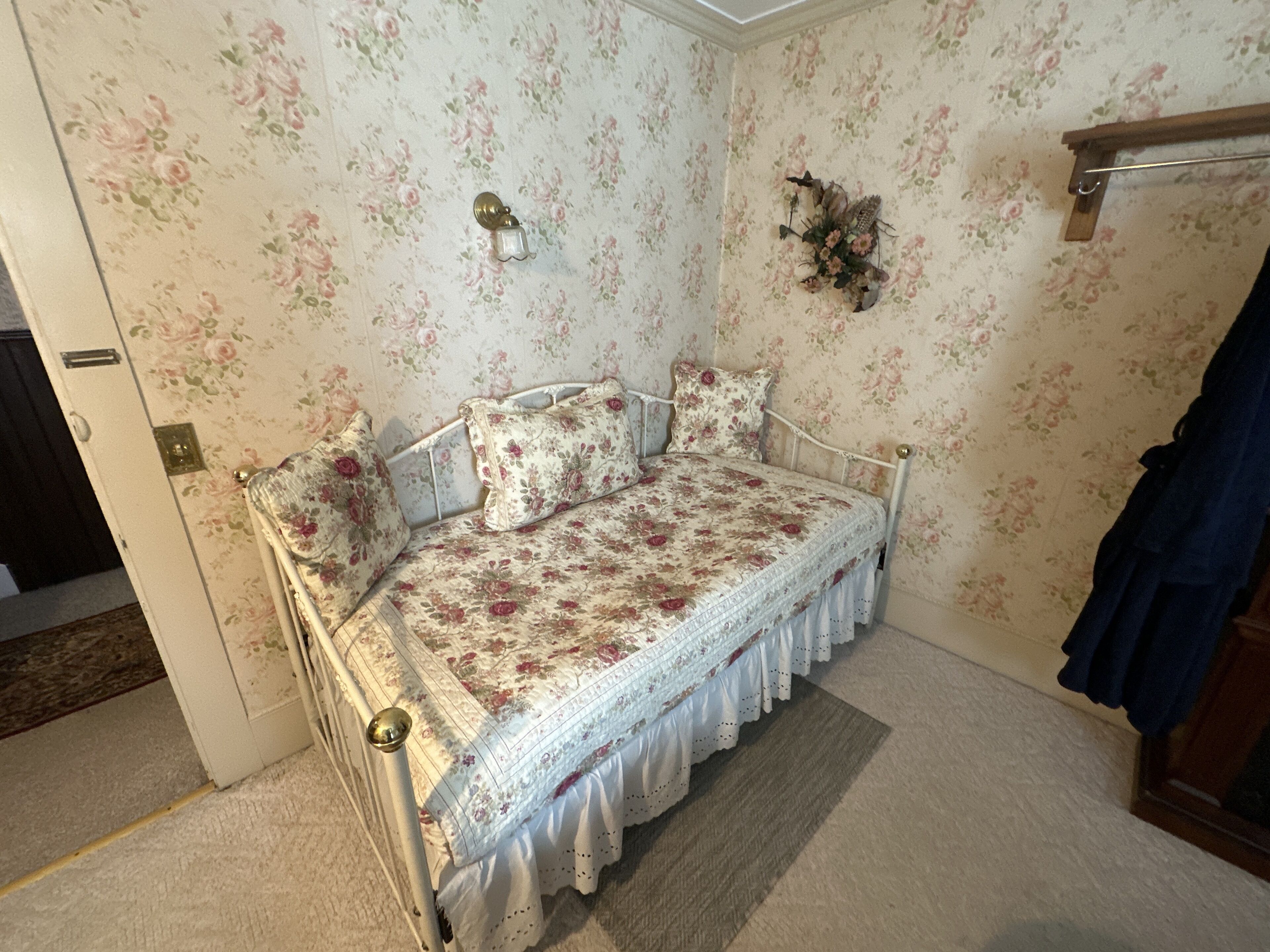 A small, cozy bedroom with floral wallpaper, a single bed with a white frame, and a wooden door.