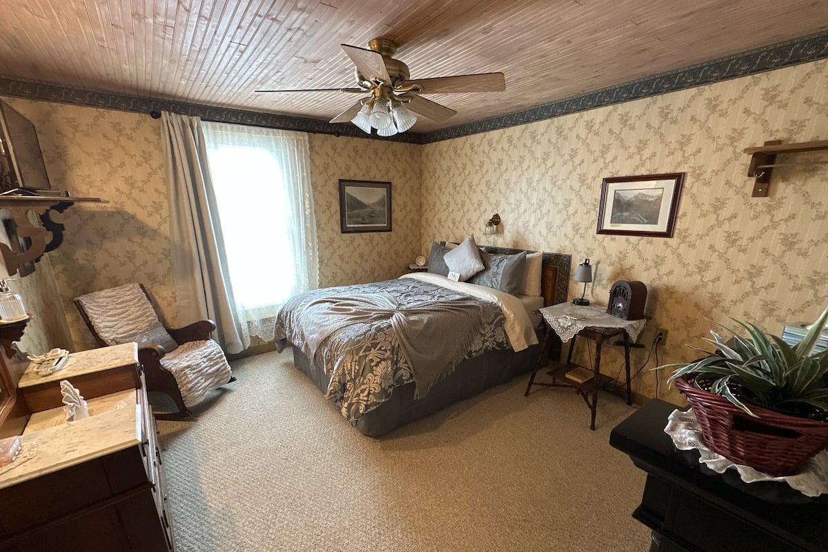 A bedroom with a bed, a desk, a chair, a ceiling fan, and a window.