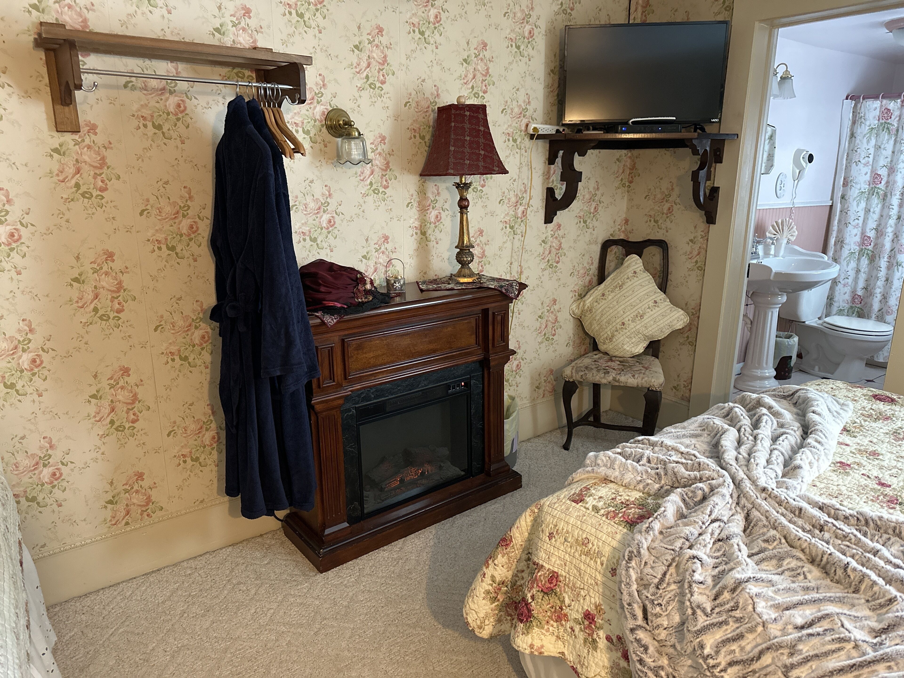 Un dormitorio con chimenea, una cama con edredón floral, una silla y un televisor.