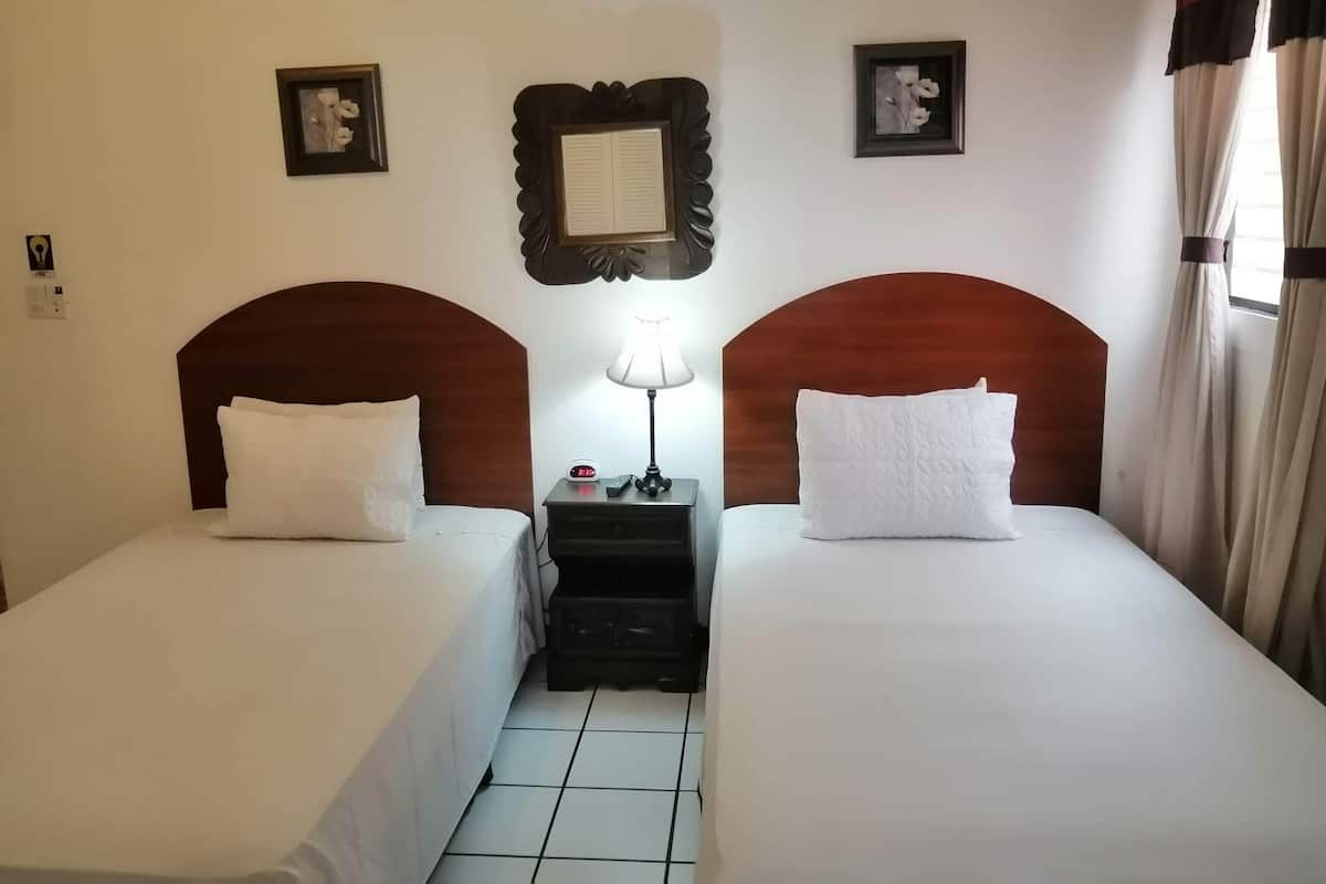 Une chambre d’hôtel avec deux lits simples, une table de chevet avec une lampe, une horloge et des tableaux encadrés au mur.