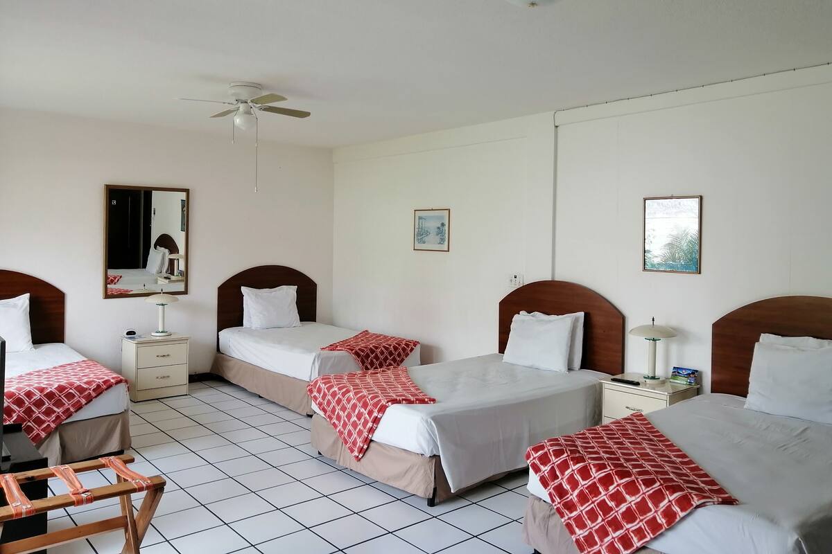 Une chambre d’hôtel avec trois lits, un ventilateur de plafond, un miroir et deux tableaux accrochés au mur.