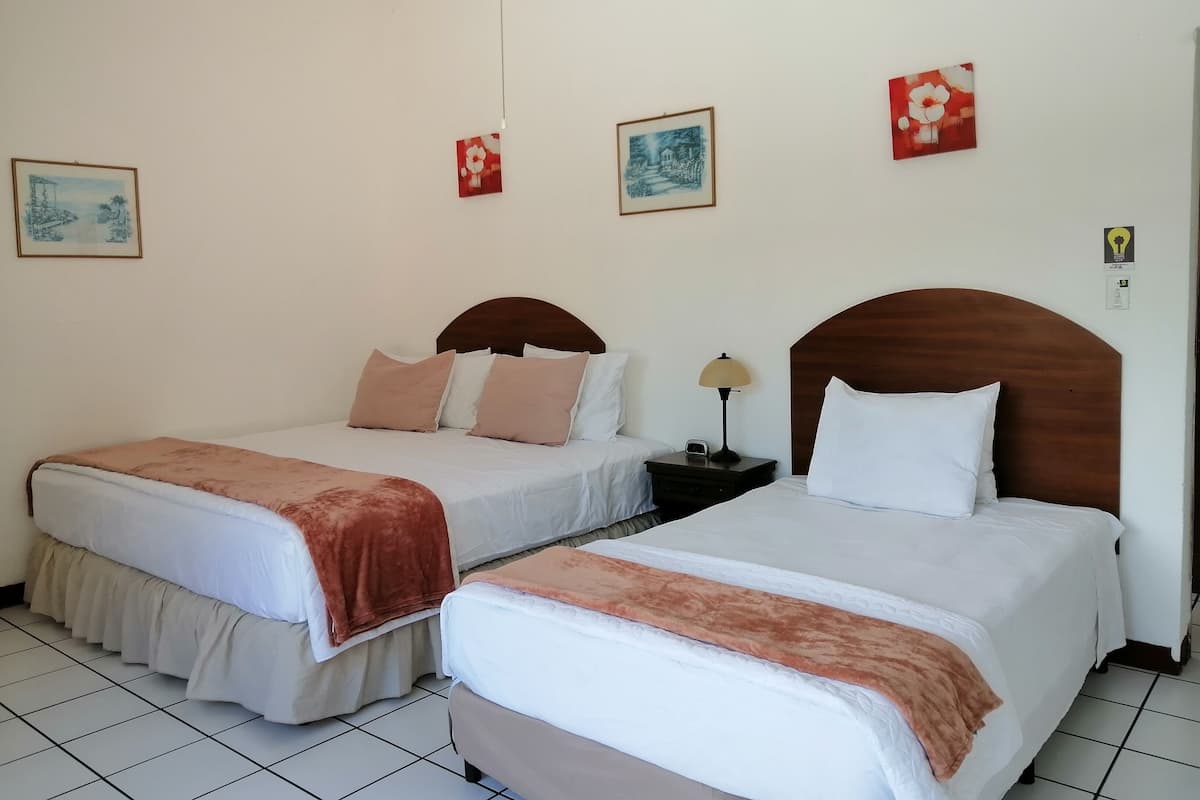 Une chambre d’hôtel avec deux lits, chacun doté d’une tête de lit en bois, de draps blancs et d’un plaid marron. Il y a deux tables de chevet avec des lampes, et les murs sont décorés d’œuvres d’art encadrées.