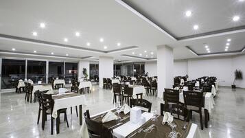 Un vaste espace dédié à la salle à manger, avec de nombreuses tables et chaises disposées en rangées, un plafond au design moderne et de grandes fenêtres à l’arrière-plan.