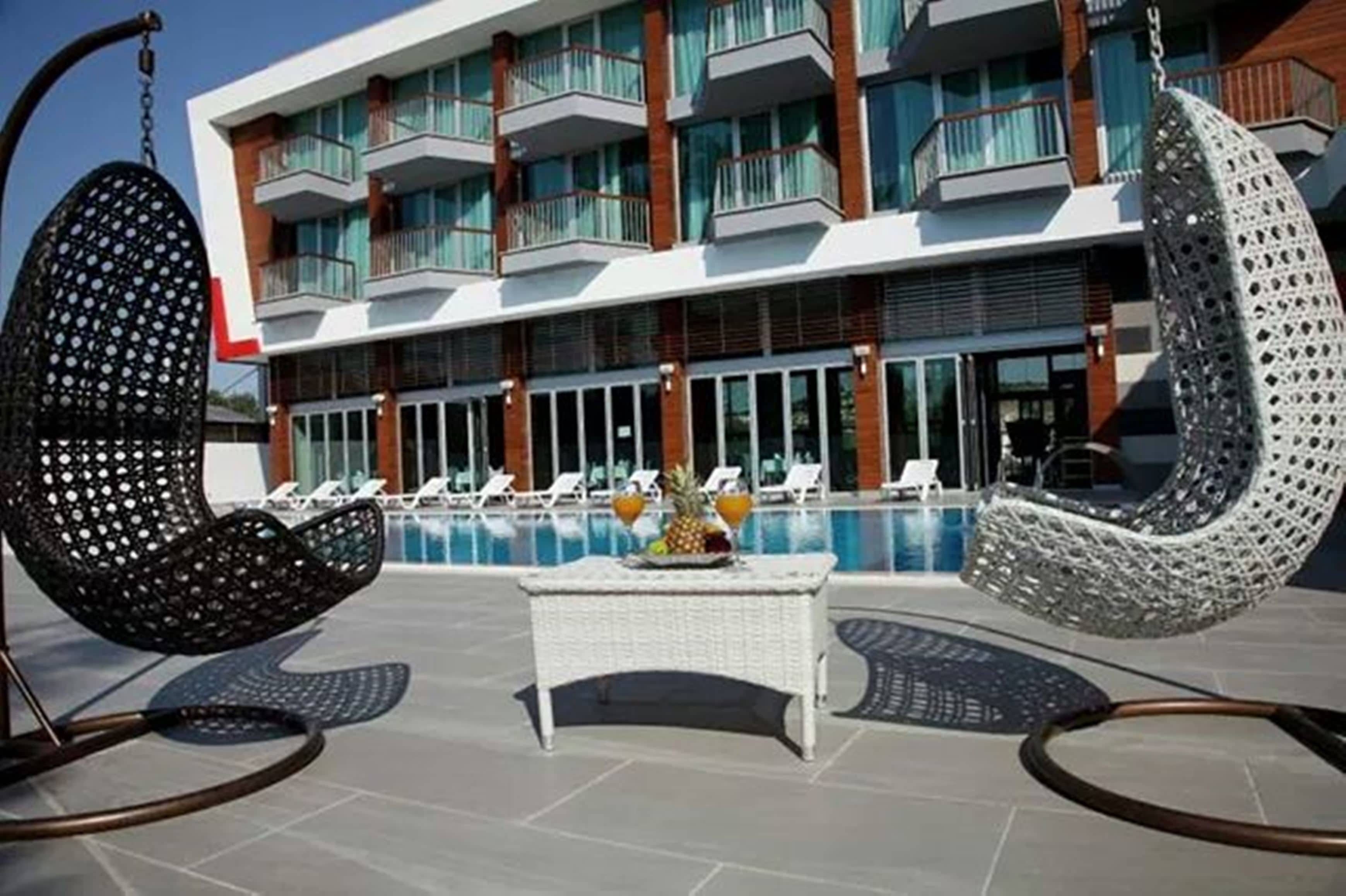 Un espace aménagé au bord de la piscine, avec deux fauteuils suspendus, une table garnie de fruits et, en arrière-plan, un bâtiment moderne doté de balcons.