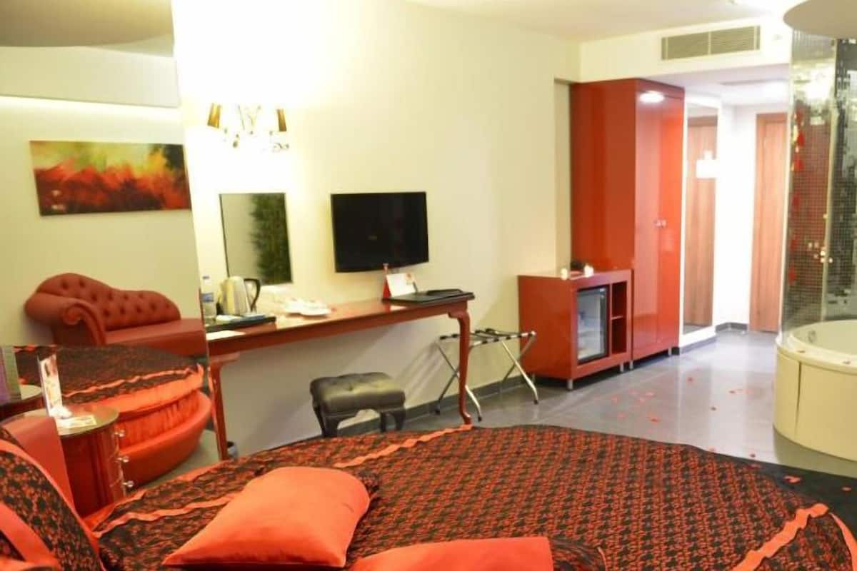 A hotel room with a bed, a desk, a television, a red sofa, a red armchair, a red ottoman, a large mirror, a wall-mounted TV, a glass shower enclosure, and a red carpet.