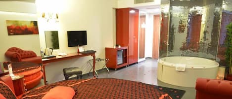 A hotel room with a bed, a desk, a television, a red sofa, a red armchair, a red ottoman, a large mirror, a wall-mounted TV, a glass shower enclosure, and a red carpet.