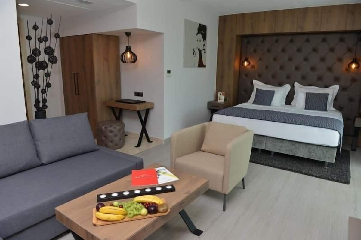 A modern hotel room with a sofa, armchair, and a bed with a tufted headboard. There is a small table with fruits and a book, and a decorative wall piece.