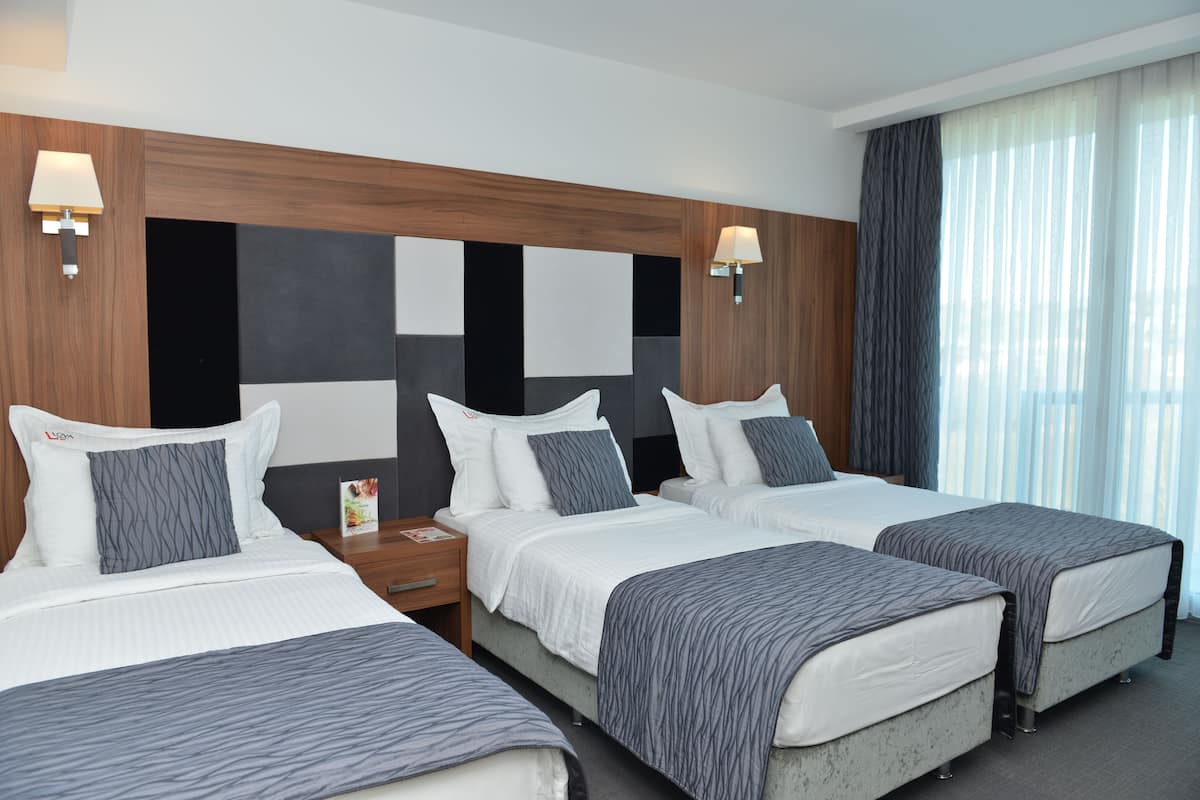A hotel room with three beds, each with a grey and white striped bedspread, wooden headboard, and bedside lamps.