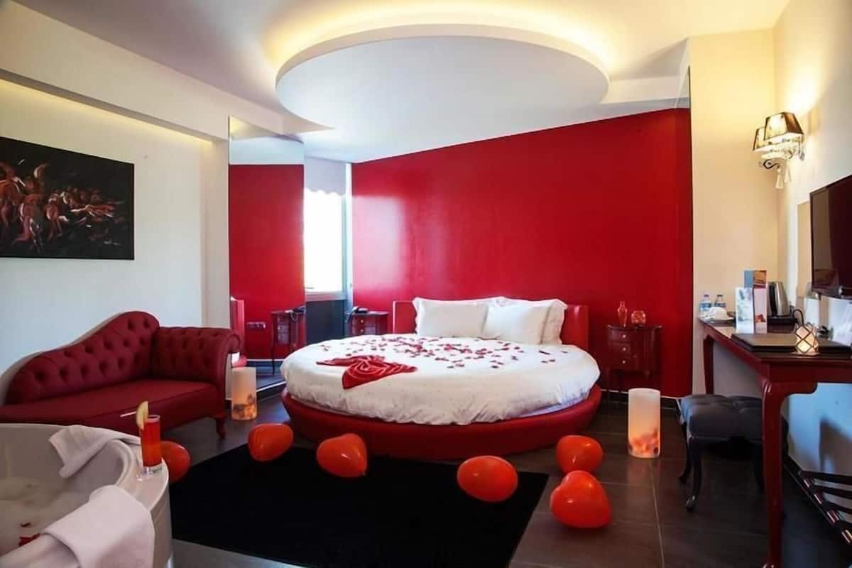 A hotel room with a red and white color scheme, a round bed, a red sofa, a desk, and a painting on the wall.