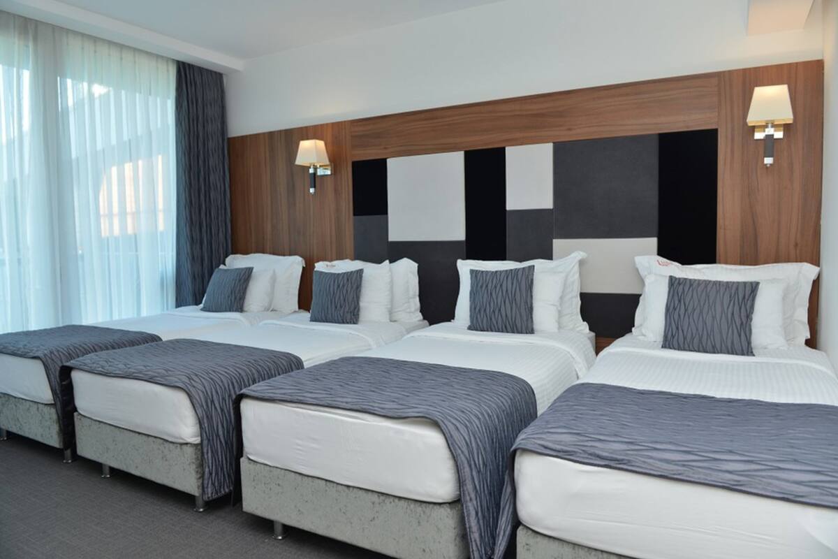 A hotel room with four beds, each with a grey blanket, white bedding, and a wooden headboard. There are two wall-mounted lamps on either side of the headboard.