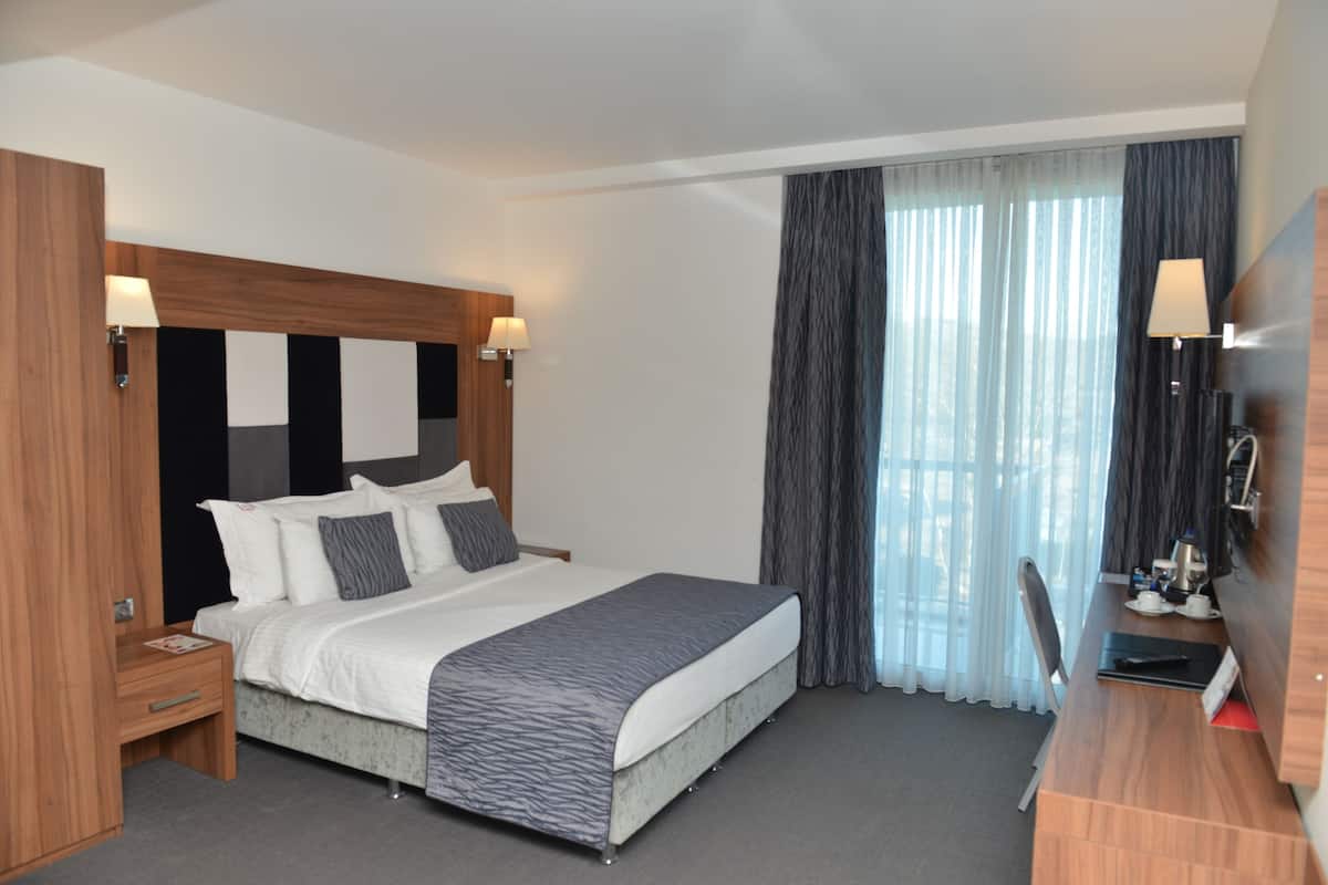A modern hotel room with a large bed, bedside lamps, a desk with a chair, and a view of the outdoors through sheer curtains.