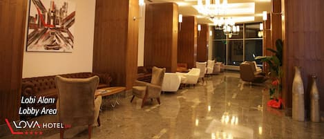 A lobby area with sofas, armchairs, and a chandelier. There is a painting on the wall and a plant on the right.