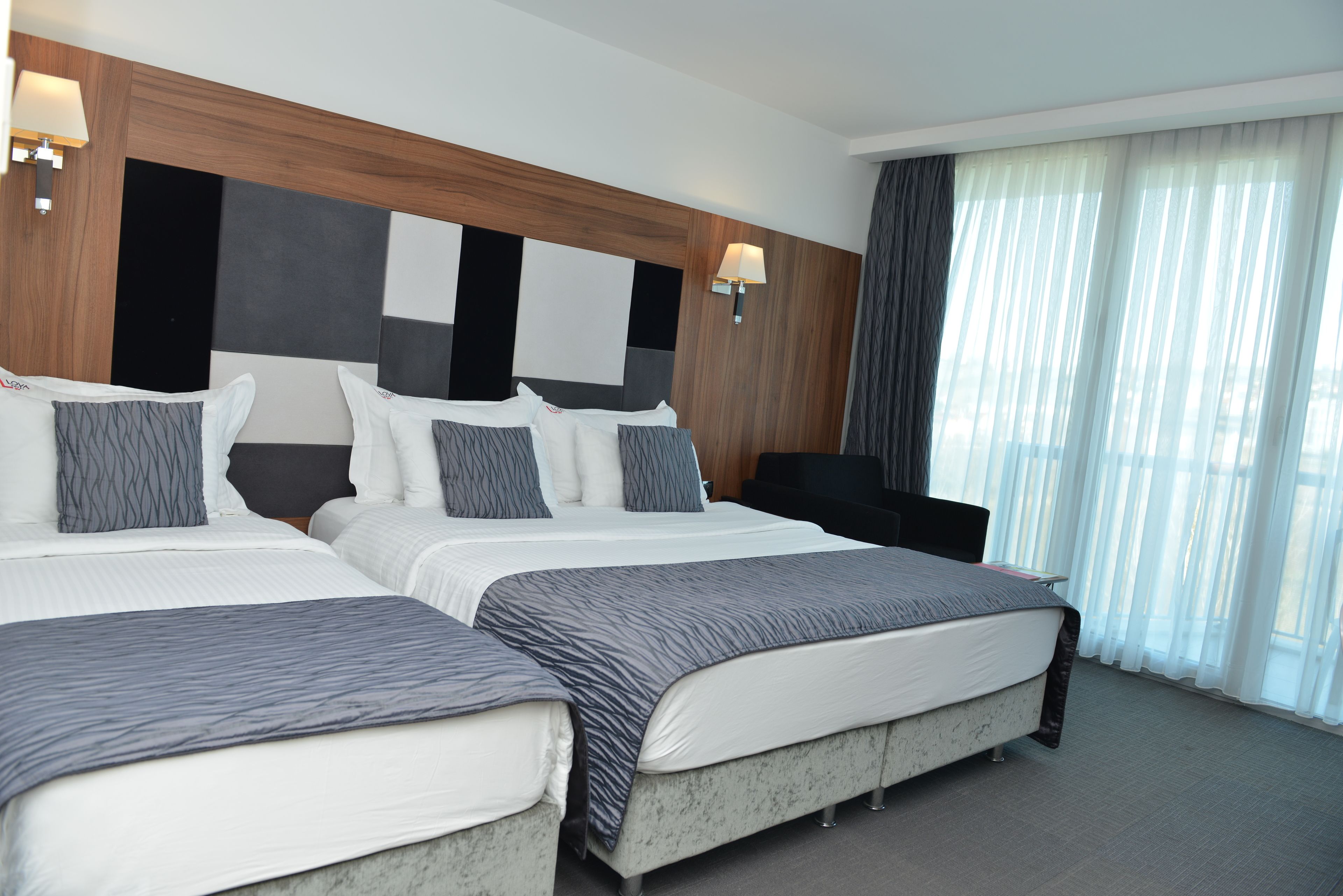 A hotel room with two beds, a large window with sheer curtains, and a wall-mounted TV.