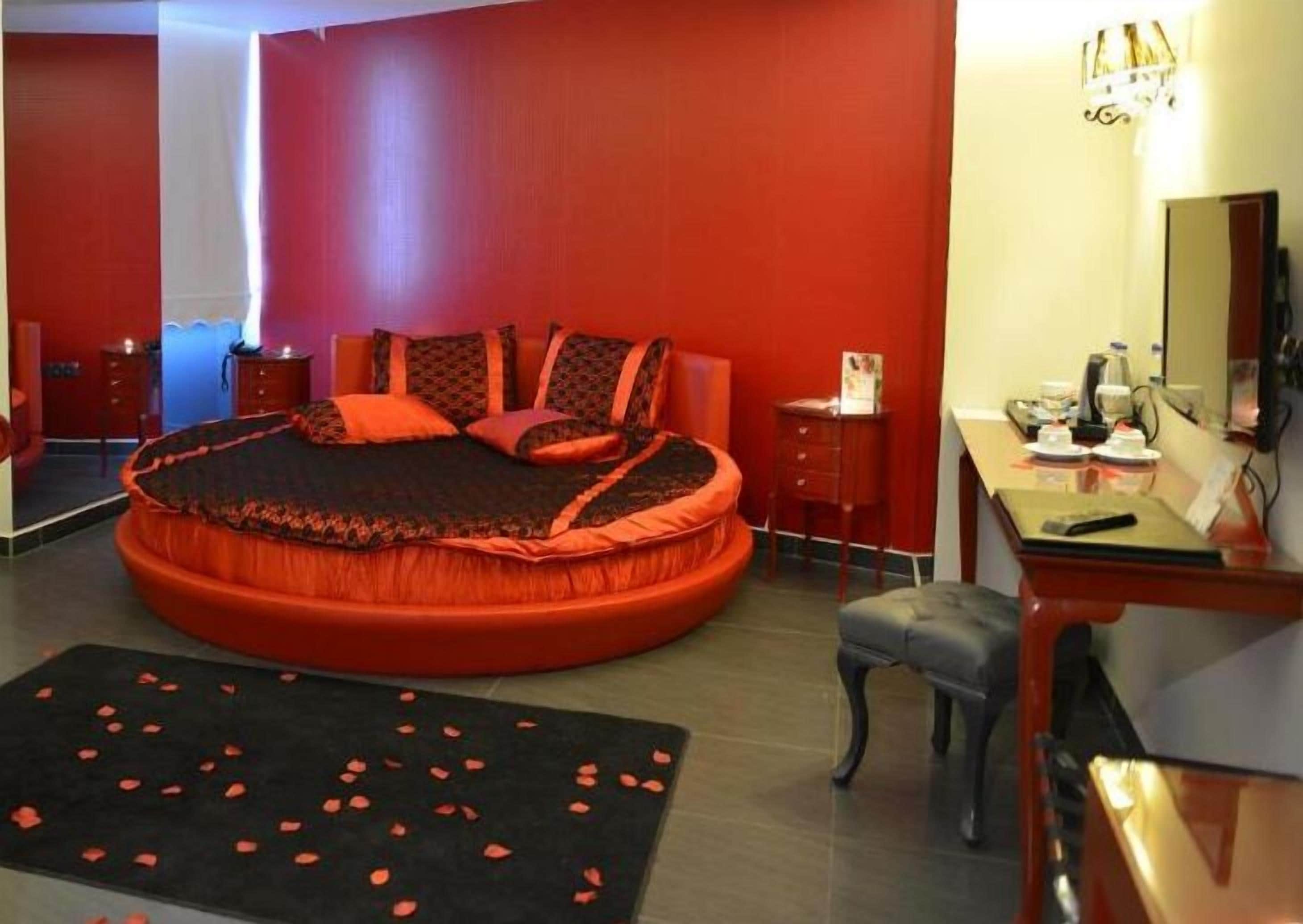 A circular bed with a red and black bedspread, a small bedside table, a mirror, and a desk with a chair.
