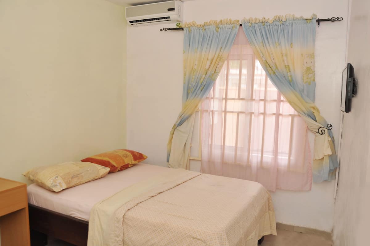 A bedroom with a bed, pillows, an air conditioner, a window with curtains, and a TV on the wall.