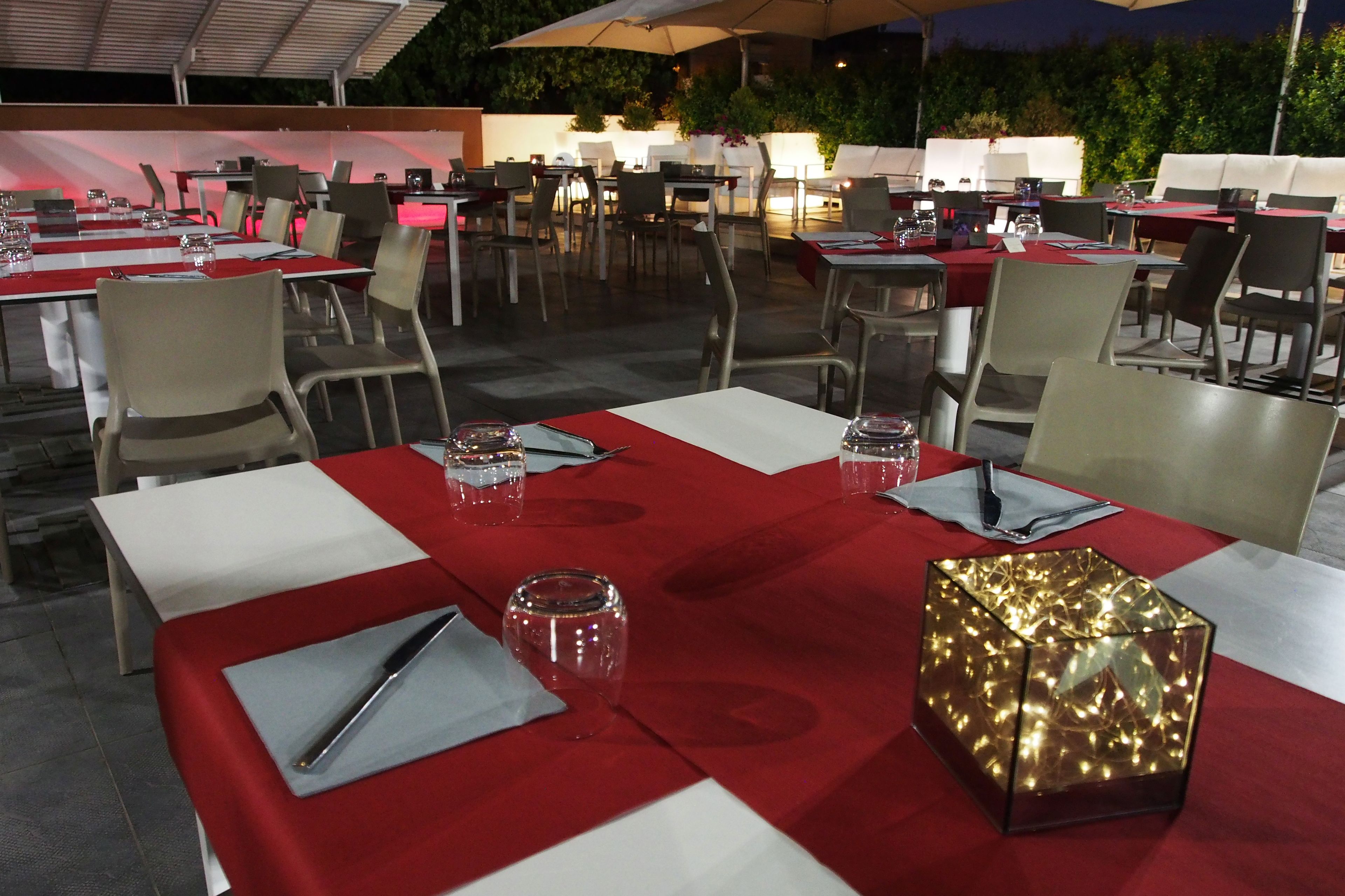 Un área de comedor al aire libre con mesas dispuestas para dos, con manteles rojos y vasos transparentes.