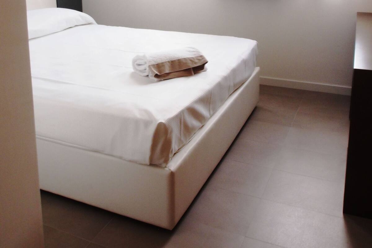 A neatly made bed with a white comforter and a folded towel, a dark patterned headboard, and a window with sheer white curtains and a dark drape.