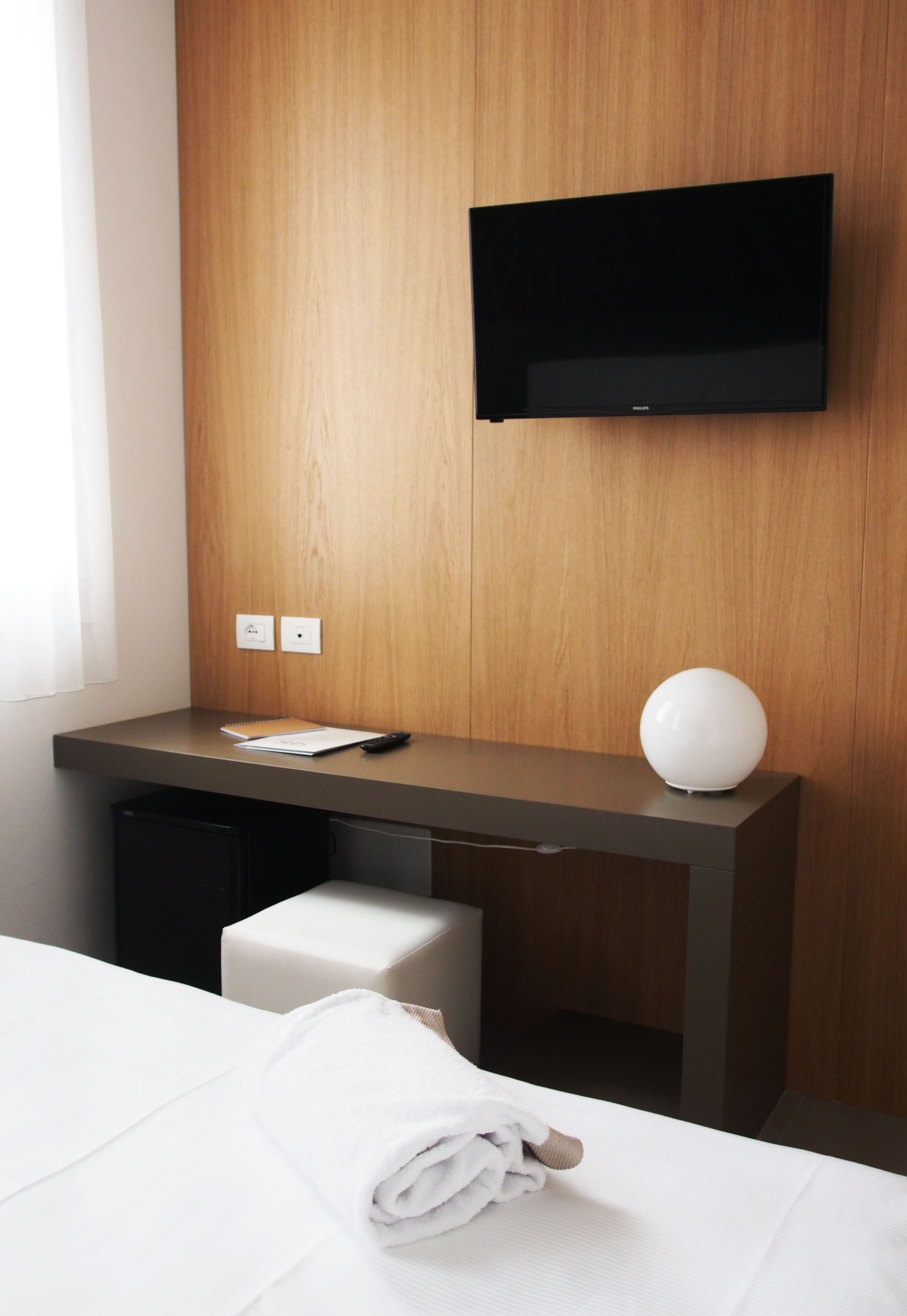 A hotel room with a wooden desk, a flat-screen TV, a white towel, and a small white stool.