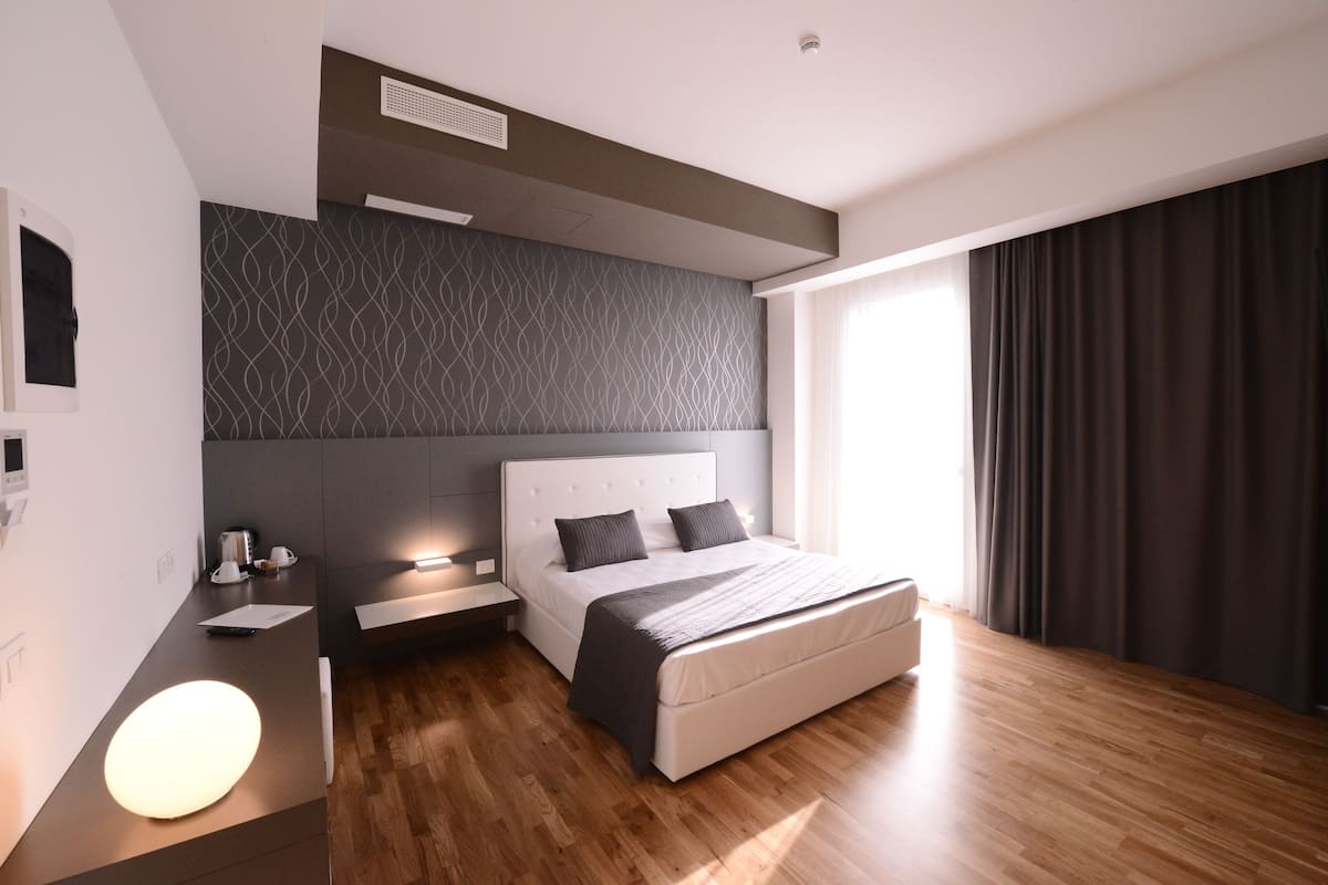 A modern bedroom with a large bed, bedside tables, a nightstand, a wall-mounted heater, and a large window with curtains.