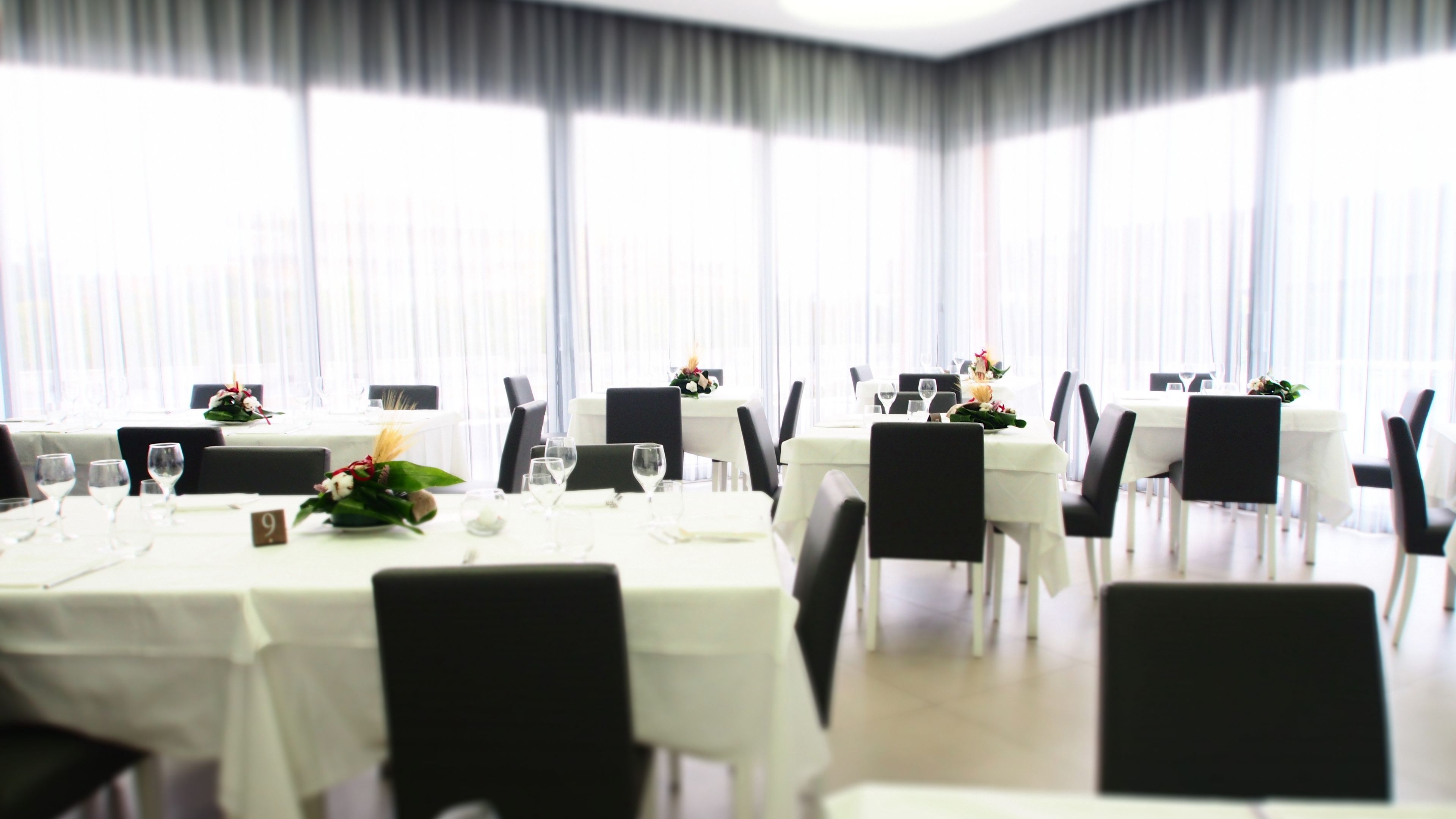 Zona pranzo con tavoli apparecchiati, sedie e un'ampia finestra con tende.