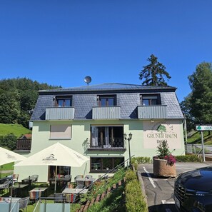 Um edifício de dois andares com uma placa que diz "Gruner Baum Hotel". Há mesas e cadeiras ao ar livre sob guarda-sóis e um carro estacionado em frente.