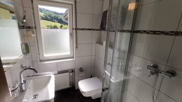 Un baño con lavamanos, inodoro y ducha blancos. Una ventana ofrece vistas a un paisaje verde.