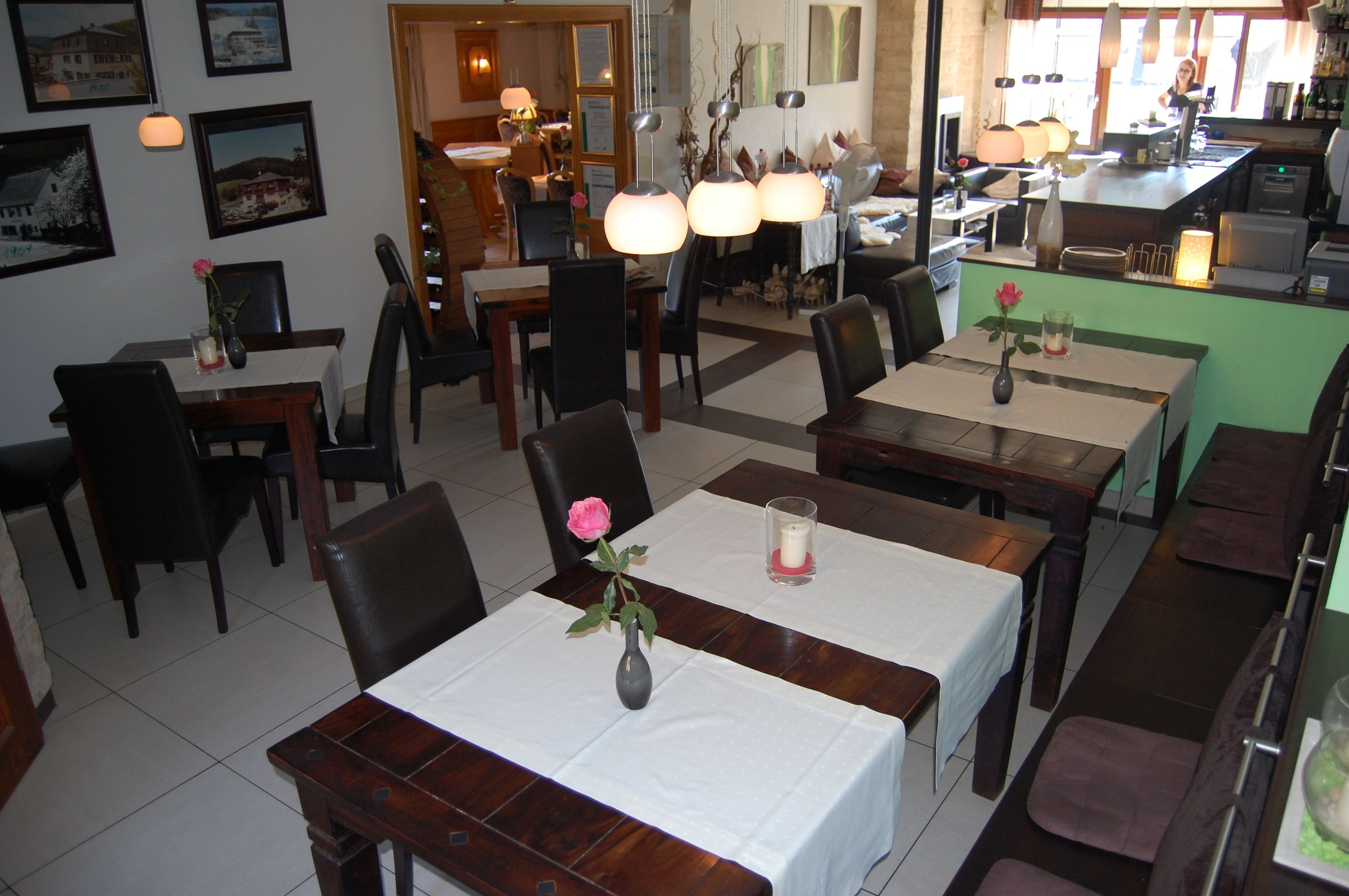 Un restaurante con varias mesas y sillas, algunas decoradas con flores y velas. Se observan cuadros en las paredes y una zona de cocina al fondo.