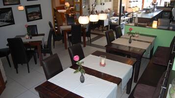 Un restaurante con varias mesas y sillas, algunas decoradas con flores y velas. Se observan cuadros en las paredes y una zona de cocina al fondo.
