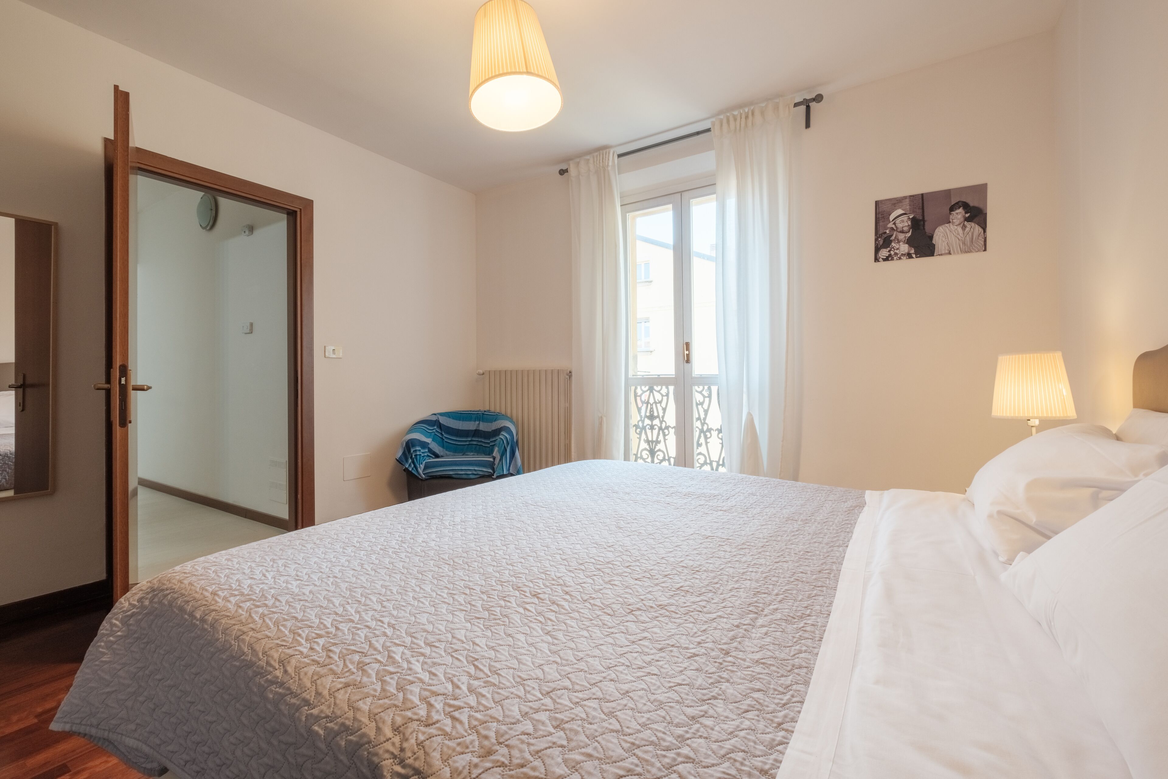 A hotel room with a large bed, a chair, a window with curtains, a wall-mounted picture, and a door leading to another room.