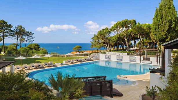 A swimming pool with lounge chairs and umbrellas, surrounded by trees and a view of the sea. - Pine Cliffs Ocean Suites, a Luxury Collection Resort & Spa (Albufeira)
