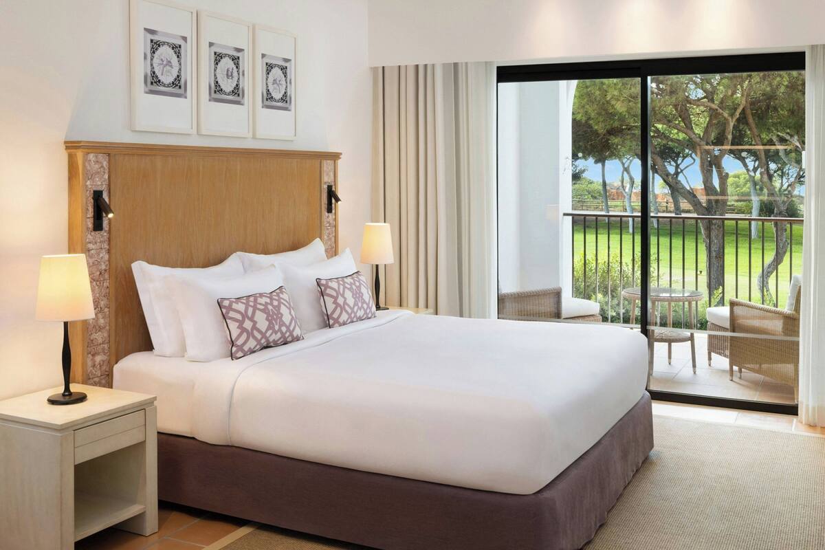 Une chambre d’hôtel avec un grand lit, deux lampes de chevet, une tête de lit en bois et une vue sur un balcon aménagé avec du mobilier d’extérieur.