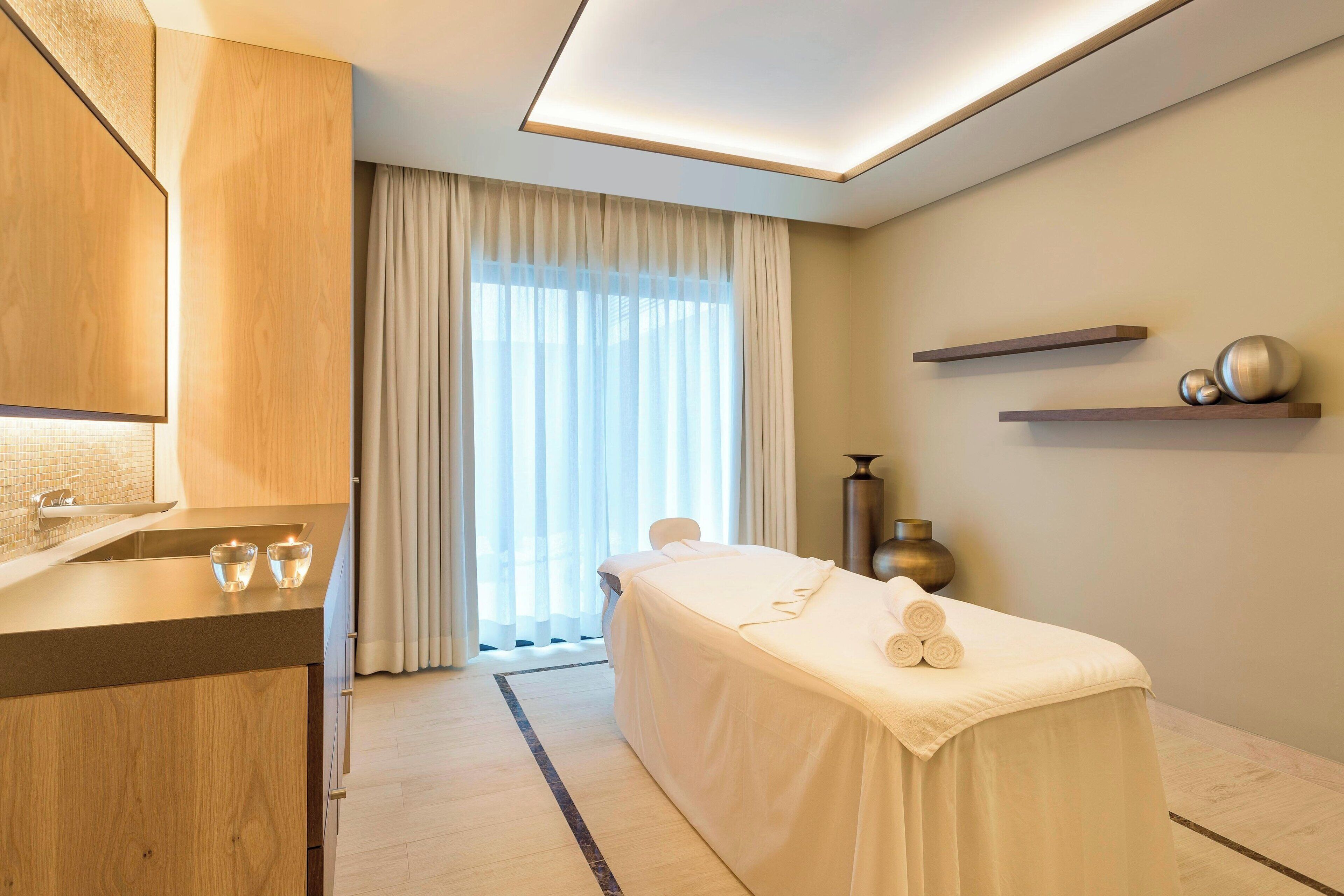 A modern spa room with a massage table, wooden cabinets, and a large window with sheer curtains.