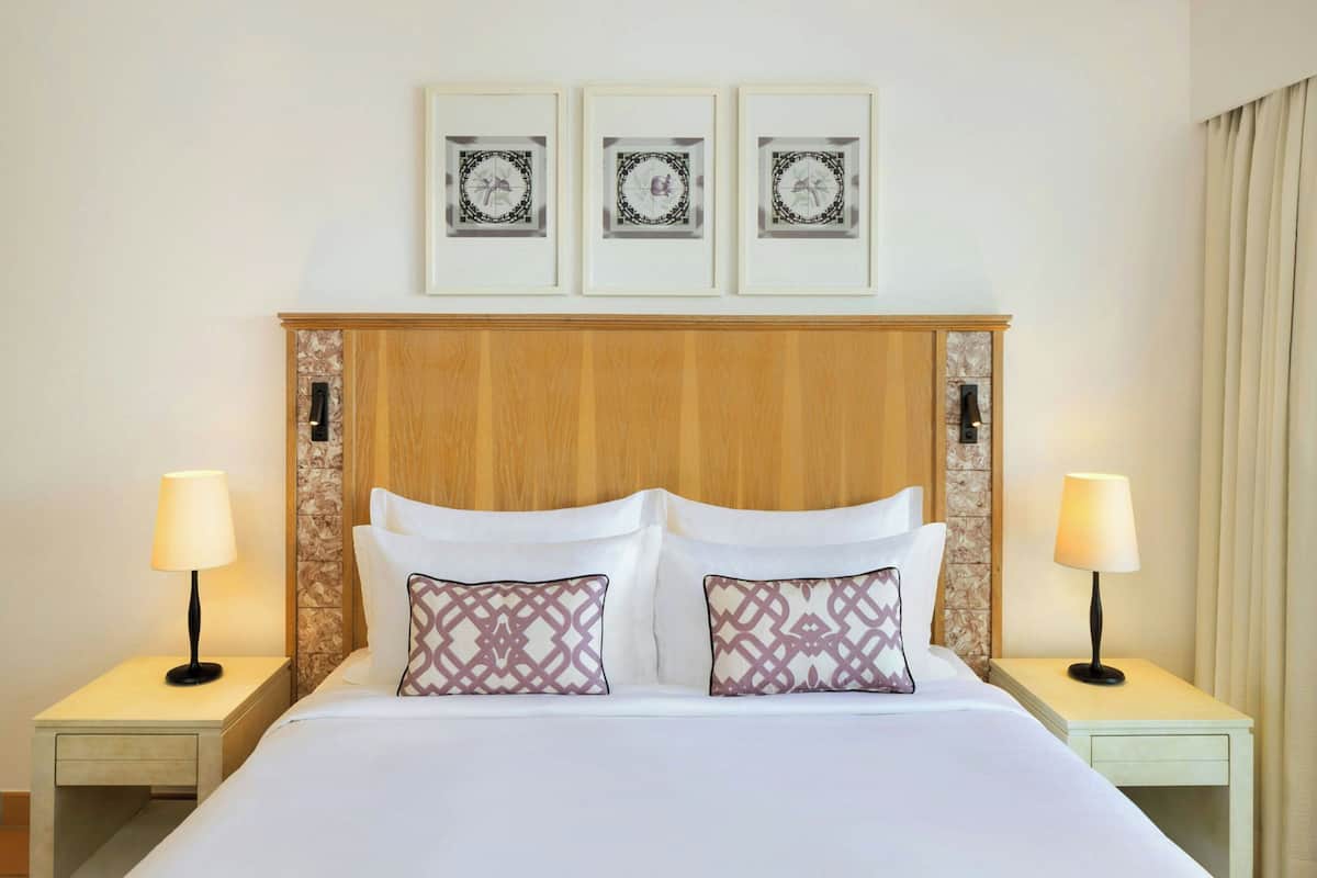 Une chambre d’hôtel avec une tête de lit en bois, deux tables de chevet avec des lampes, un lit avec du linge de lit blanc et des oreillers à motifs, et trois tableaux encadrés accrochés au mur.