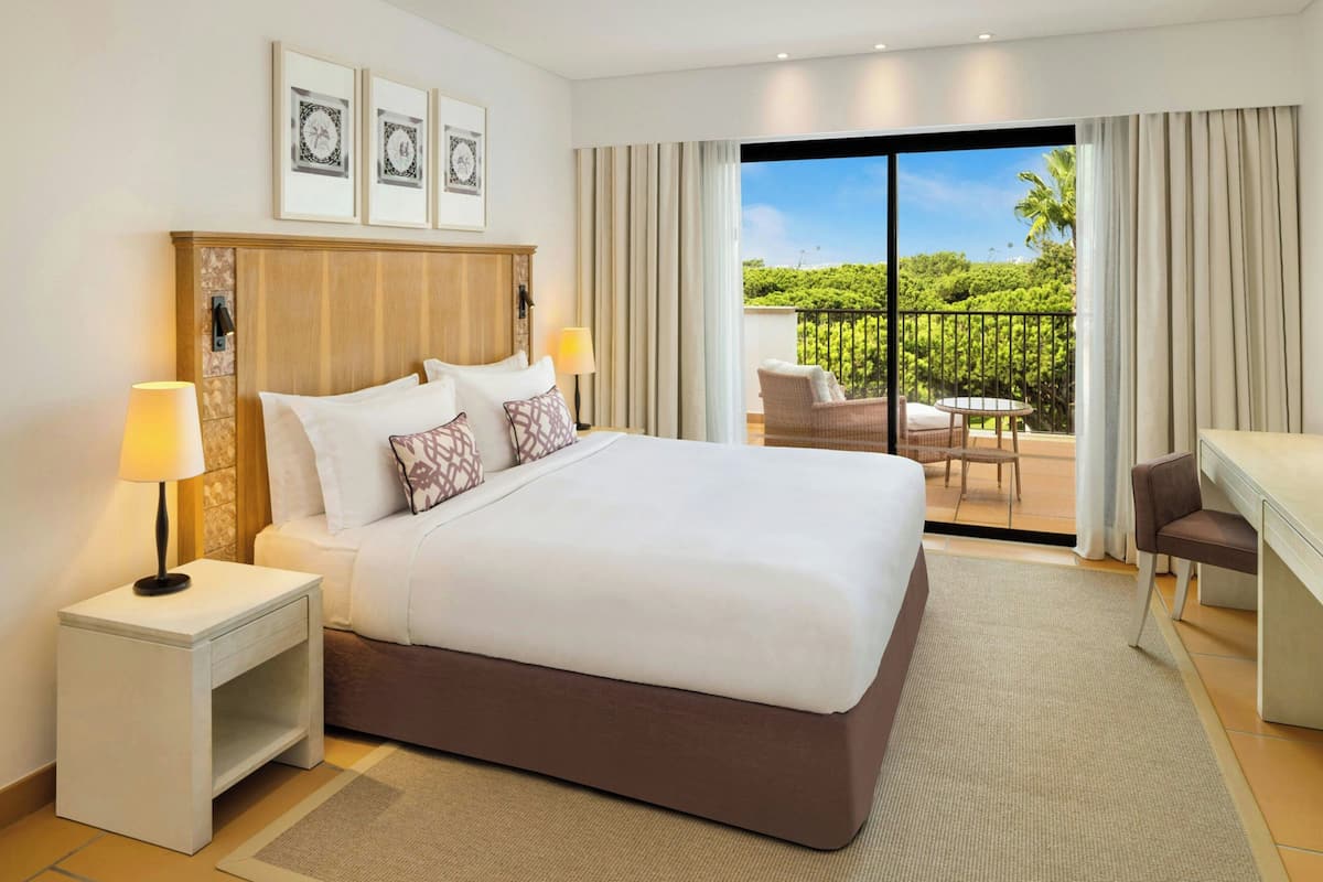 Une chambre d’hôtel avec un grand lit, deux tables de chevet avec des lampes, un bureau, une chaise et un balcon donnant sur un paysage verdoyant.