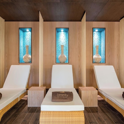 A spa room with three white lounge chairs, each with a towel labeled "Serenity." The room has wooden paneling and a mosaic tile wall with a vase.
