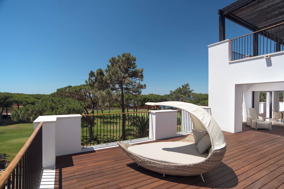 Une terrasse extérieure moderne, dotée d'un fauteuil en osier aux lignes courbes et d'un canapé blanc, offrant une vue imprenable sur un paysage verdoyant et un ciel bleu azur.