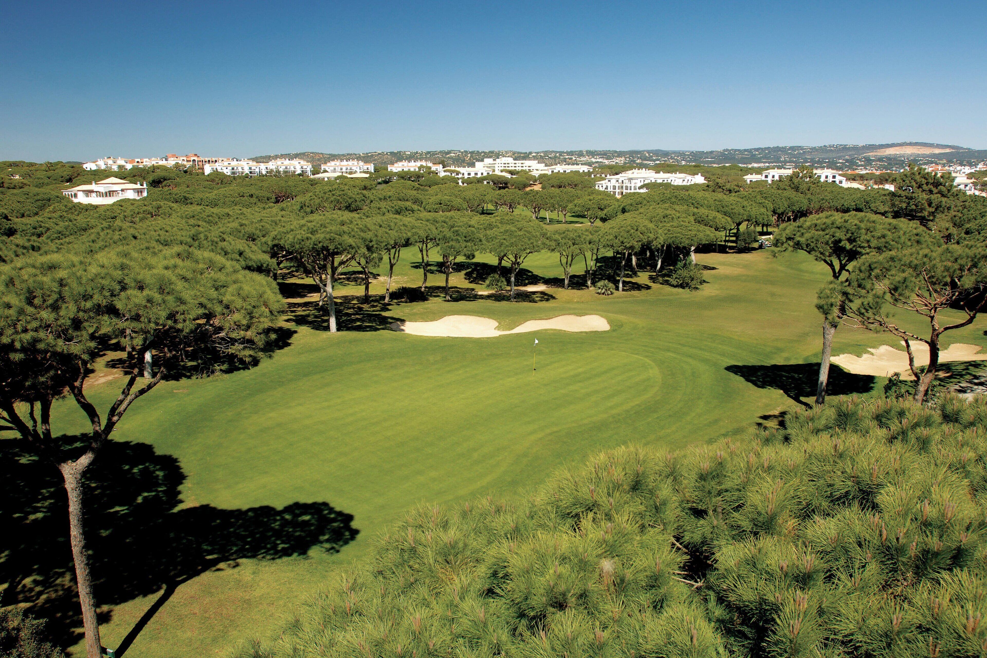 Campo de golf con búnkeres de arena, árboles y una vista distante de edificios blancos.
