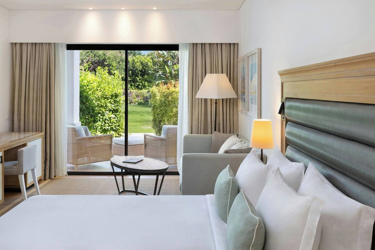Une chambre d’hôtel comprenant un lit, un bureau, une chaise, un canapé, une table et une lampe. La fenêtre offre une vue sur un jardin.