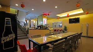 Un restaurant avec une longue table, des chaises en osier et des chaises rouges. On aperçoit un escalier au fond et un téléviseur fixé au mur.