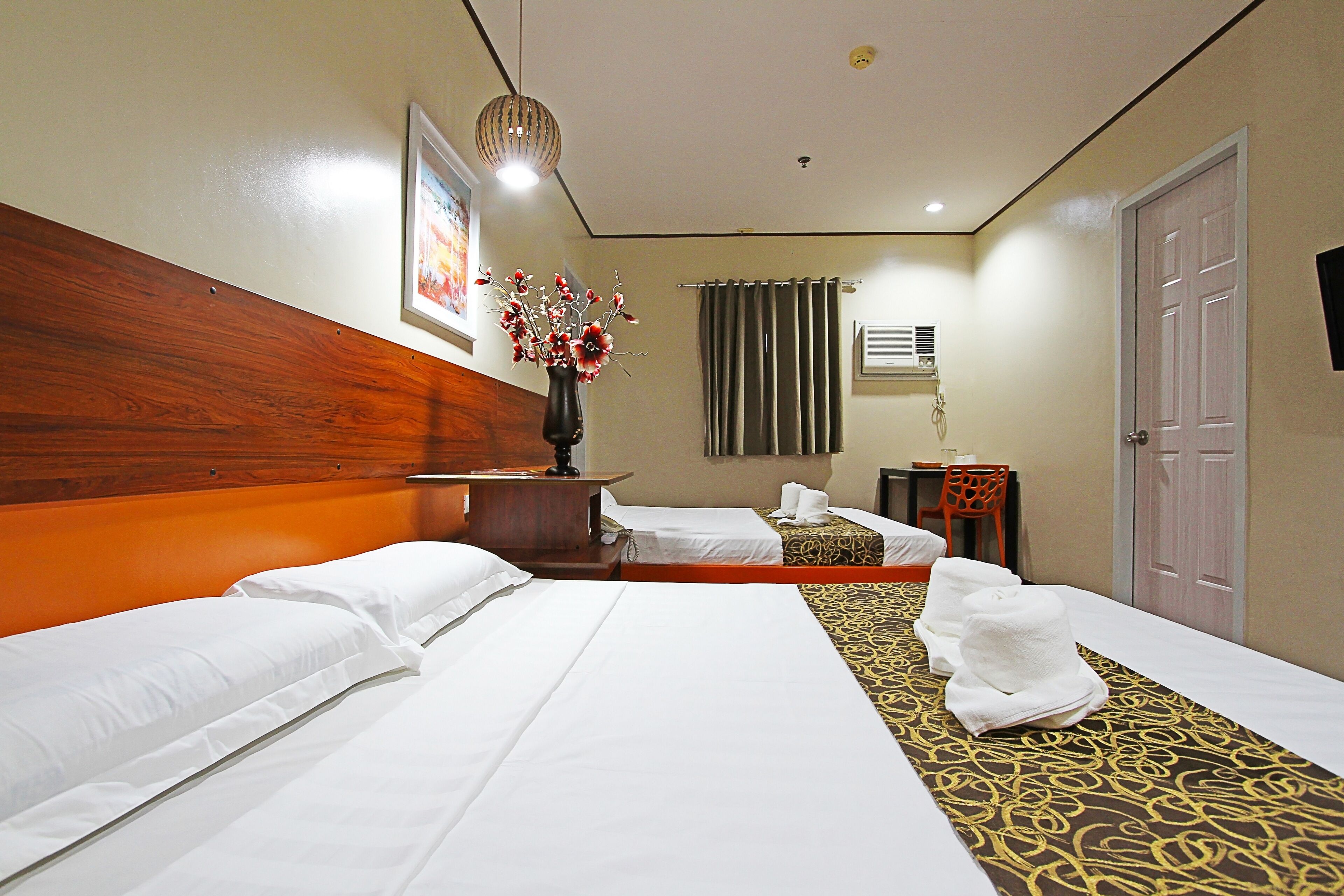 A hotel room with two beds, a wooden headboard, a vase with flowers, a desk with a chair, and a door.